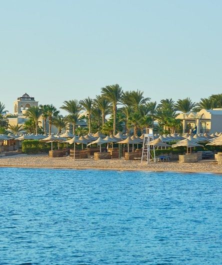 Ein Sandstrand mit zahlreichen Sonnenschirmen und Palmen vor einer Hotelanlage, aufgenommen vom blauen Meer aus.