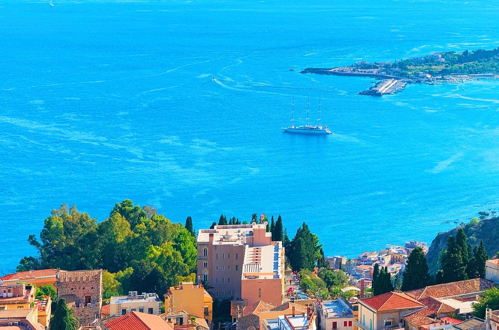 Blick von oben auf eine Küstenstadt mit rötlichen Dächern. Dahinter erstreckt sich ein azurblaues Meer, auf dem ein großer Segler ankert.