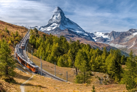 Eine orangefarbene Zahnradbahn der Gornergrat Bahn fährt auf einem steilen, kurvigen Hang in den Schweizer Alpen. Im Hintergrund erhebt sich das markante Matterhorn mit seiner schneebedeckten Spitze unter einem dramatischen Himmel mit Wolken und blauem Himmel. Die Landschaft ist von grünen Nadelbäumen und herbstlich gefärbten Gräsern geprägt, die den Gebirgshang bedecken.