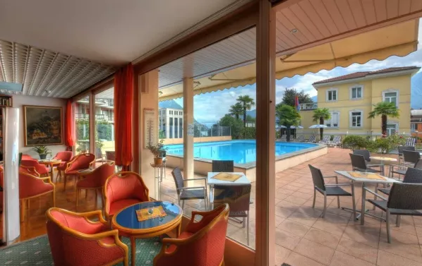 Das Innere einer Hotellobby oder eines Loungebereichs mit großen Fenstern, die den Blick auf einen Außenpool und ein hellgelbes Hotelgebäude freigeben. Im Raum befinden sich mehrere gemütliche, rote Sessel und kleine Tische. Draußen am Pool sind weitere Tische mit Sonnenschirmen und Liegestühle zu sehen, alles unter einem blauen Himmel mit einigen Wolken.